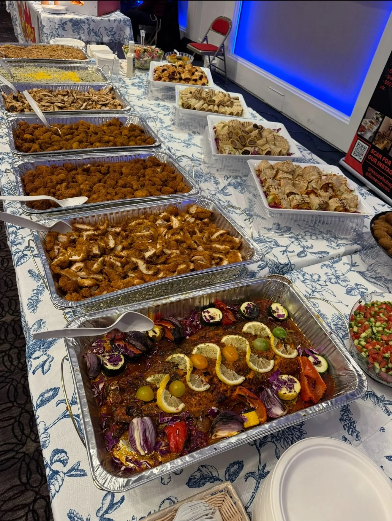 A buffet table with various dishes including fried chicken, salads, and desserts, set on a white tablecloth with a blue floral pattern.