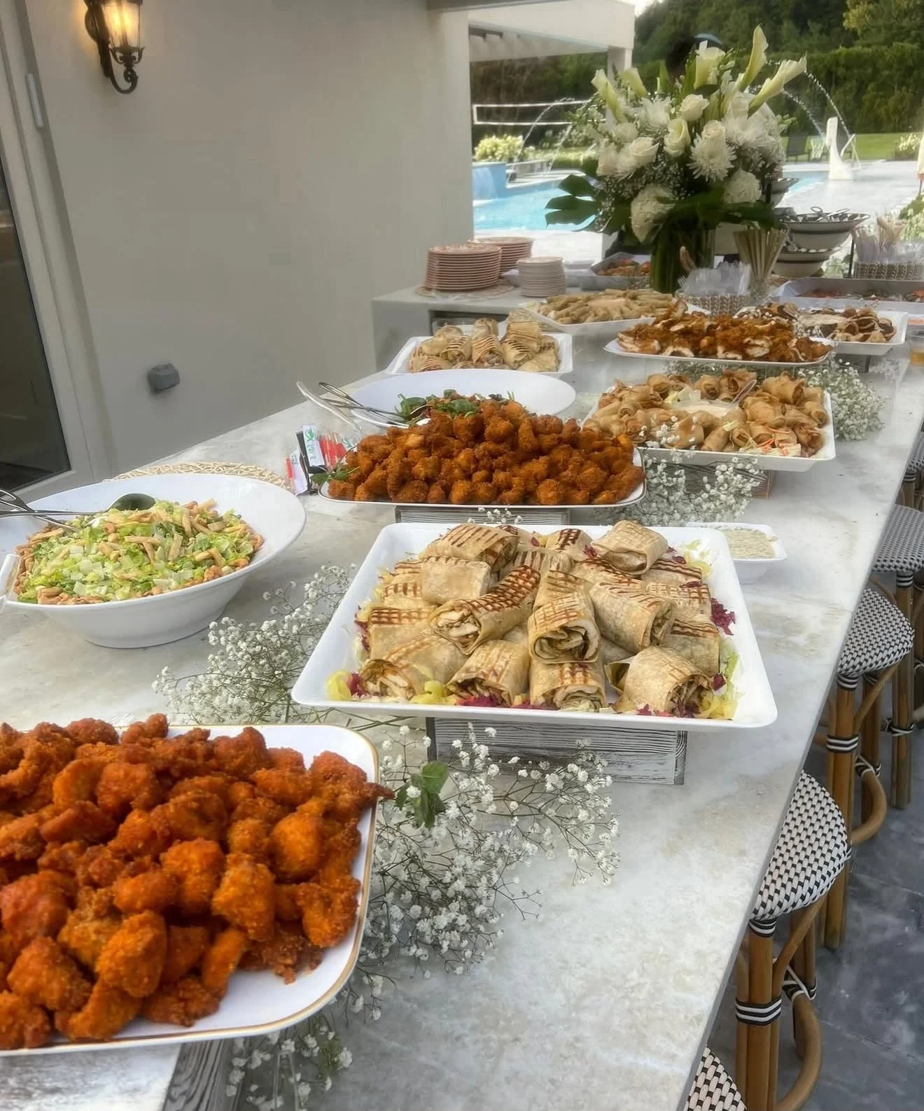 Buffet table with various dishes including spring rolls, fried chicken, and broccoli salad, adorned with white floral decorations, overlooking a pool outside.