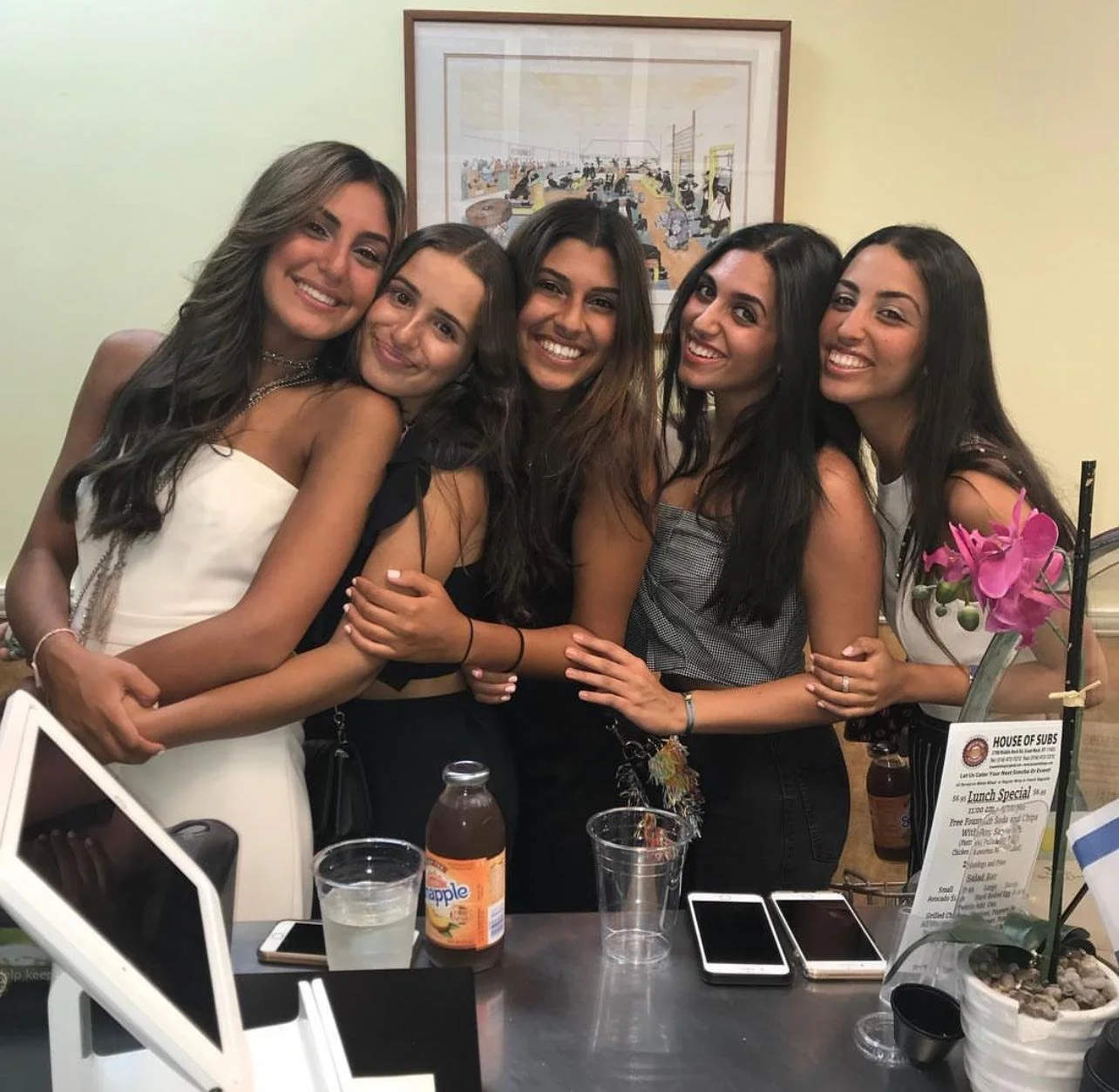 Five young women are hugging and smiling together at a social gathering in a restaurant or cafe, with drinks and smartphones on the table in front of them.