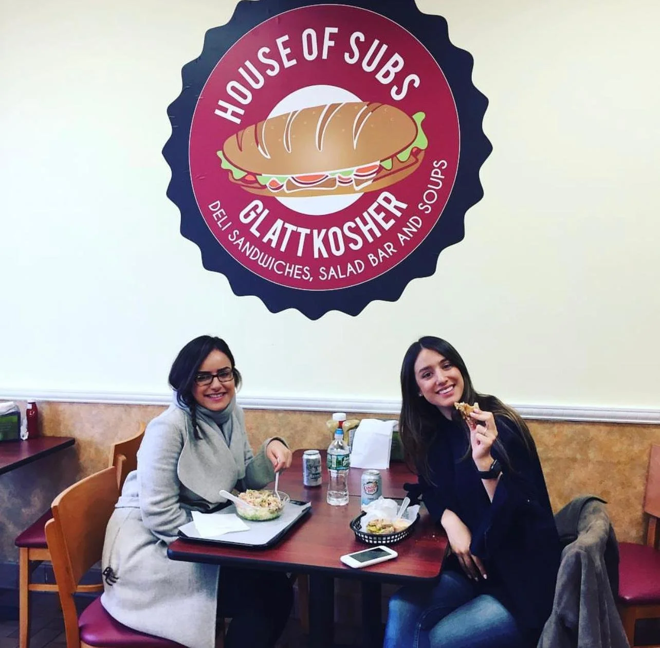 Two women sitting at a restaurant table, smiling, with food and drinks. Behind them, a large sign features a sandwich illustration and the text 'House of Subs Glatt Kosher Deli, Sandwiches, Salad Bar, and Soups.'
