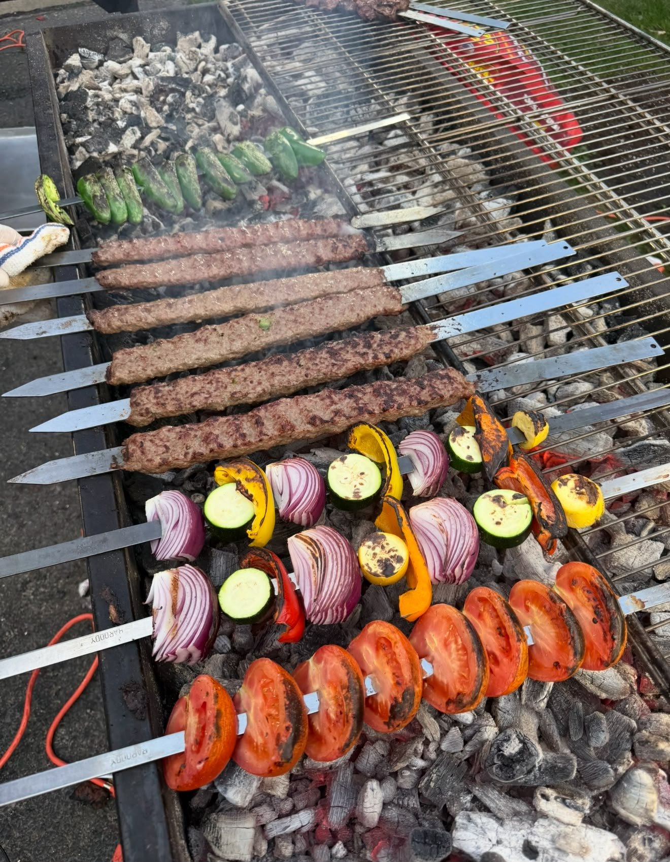 Barbecue grill with skewers of ground beef, vegetables, and tomatoes cooking over hot coals.