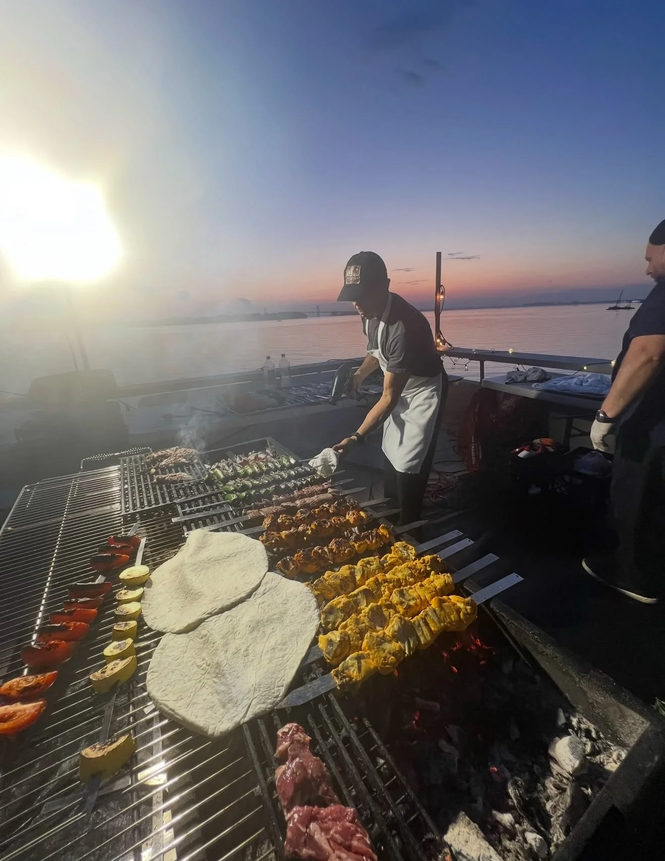 People grilling food on a barbecue grill near a body of water during sunset, with various meats and vegetables on the grill and some plates or towels nearby.
