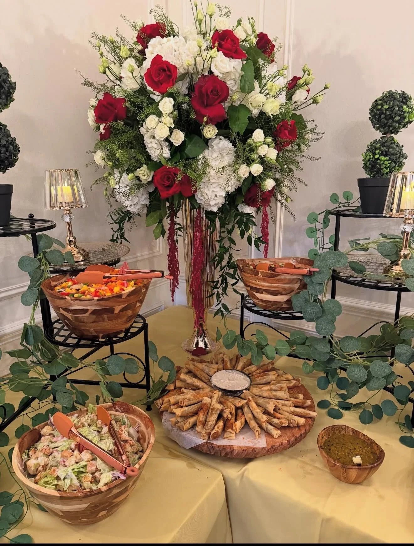 A large floral arrangement of red and white roses with green foliage, placed on a tall vase at a buffet table. Surrounding the arrangement are bowls of salad, vegetable dip, and small potted plants, with decorative lighting and greenery accents.