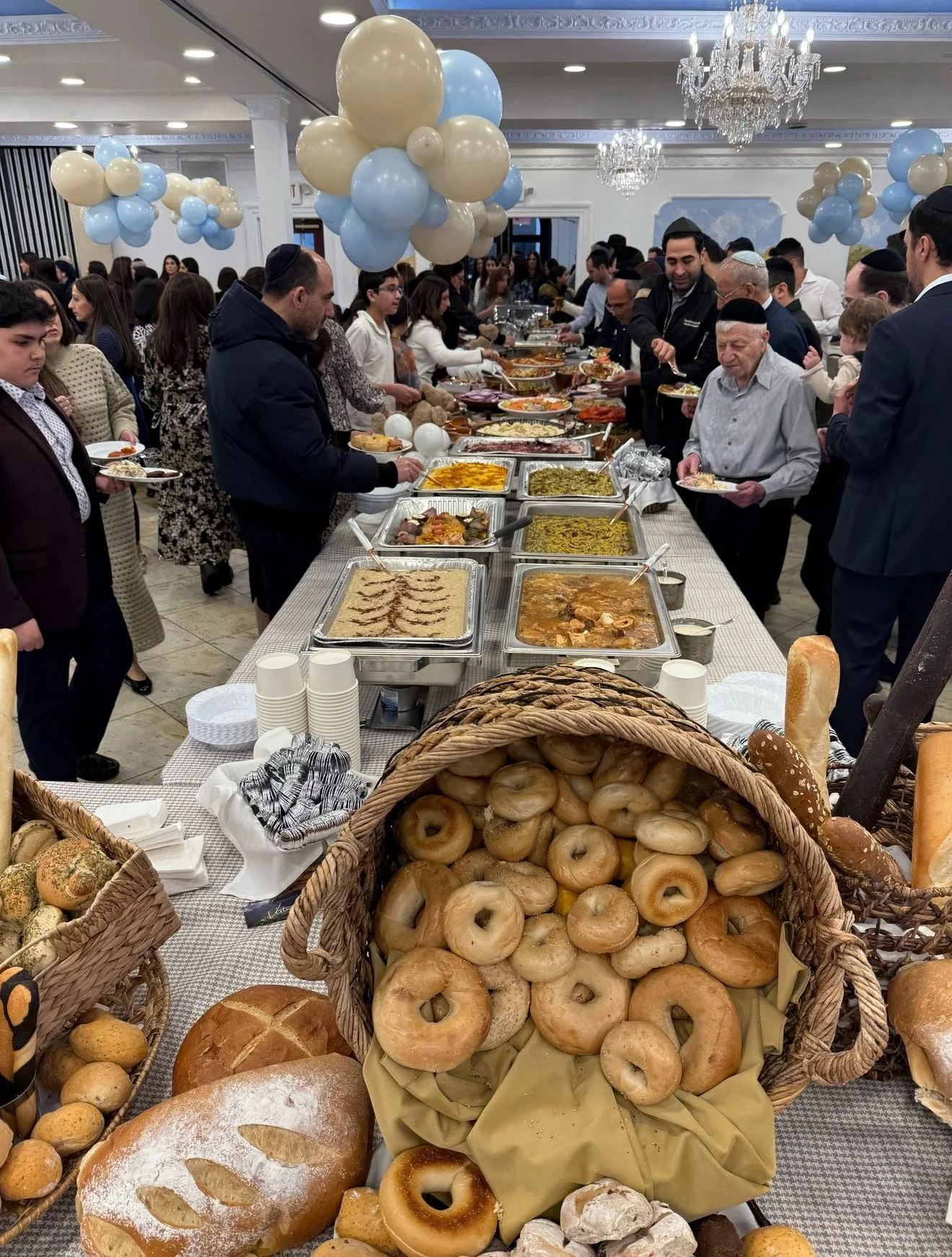 People at a buffet table with various traditional dishes, breads, and bagels, decorated with balloon arrangements and chandeliers in a banquet hall.