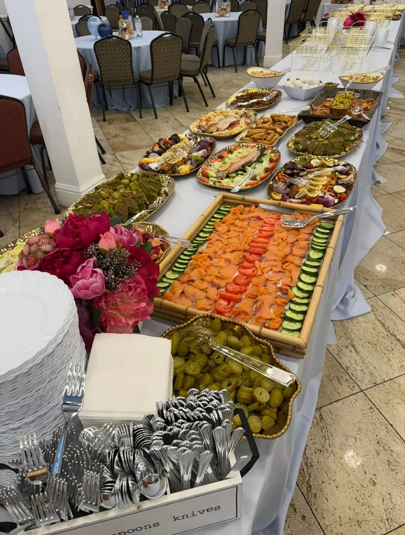 A buffet table with a variety of food including smoked salmon, vegetables, fruit, olives, and dips, with plates, napkins, and utensils nearby, set in a banquet hall with decorated tables in the background.