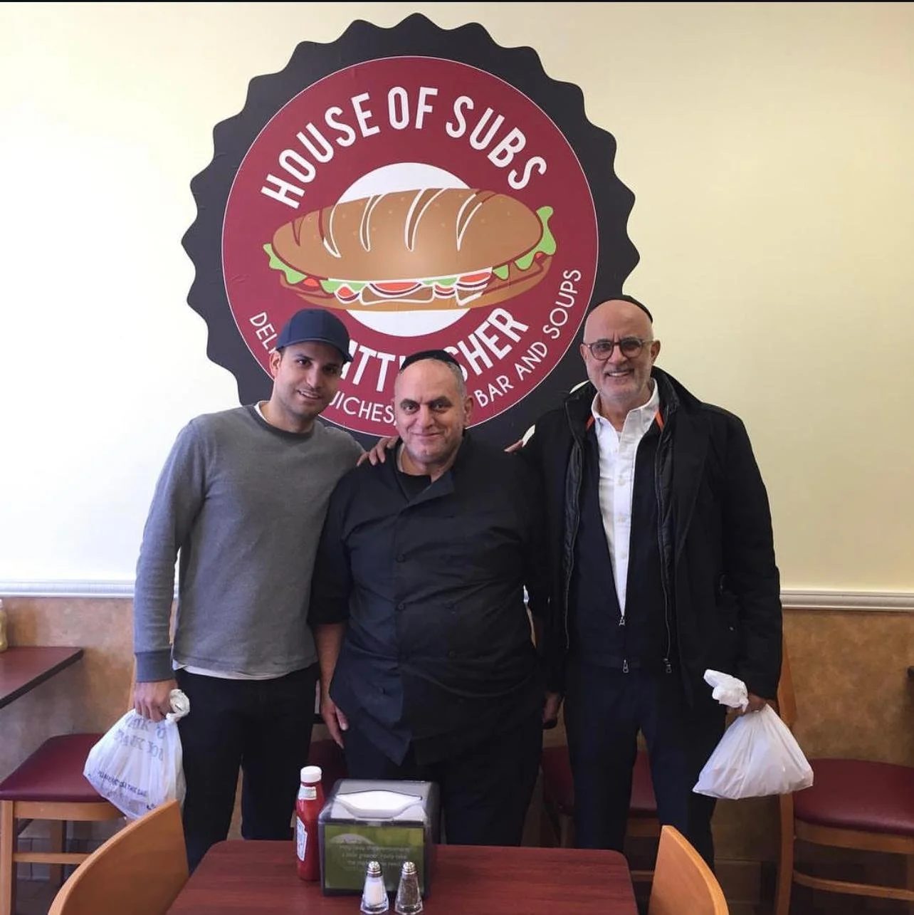 Three men standing inside a restaurant with a large logo of a sandwich behind them, smiling for the photo. The logo reads 'House of Subs' and 'Delicious Sandwiches, Bar and Soups.'