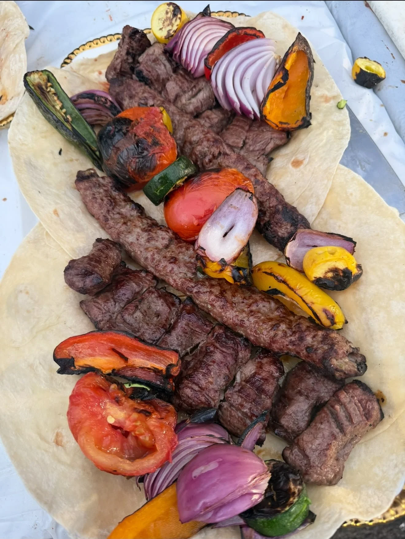 Close-up of grilled meat and vegetables on a flatbread, including slices of beef, grilled peppers, tomatoes, onions, and zucchini.