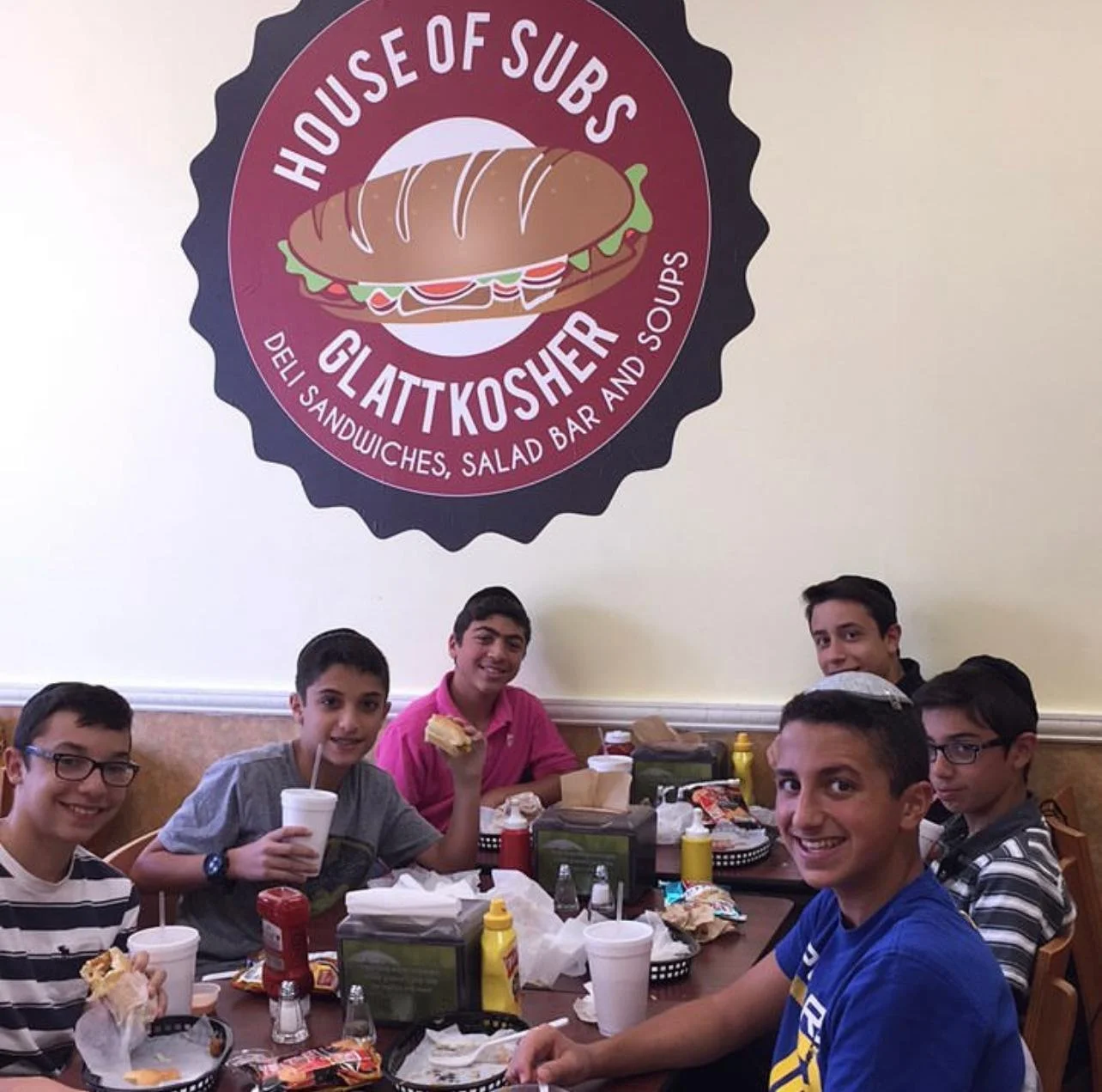 Six boys sitting at a table inside a restaurant called House of Subs, smiling and holding food and drinks, with condiments, napkins, and meal leftovers on the table and a large sign on the wall behind them reading 'House of Subs Gluten Kosher Deli, S