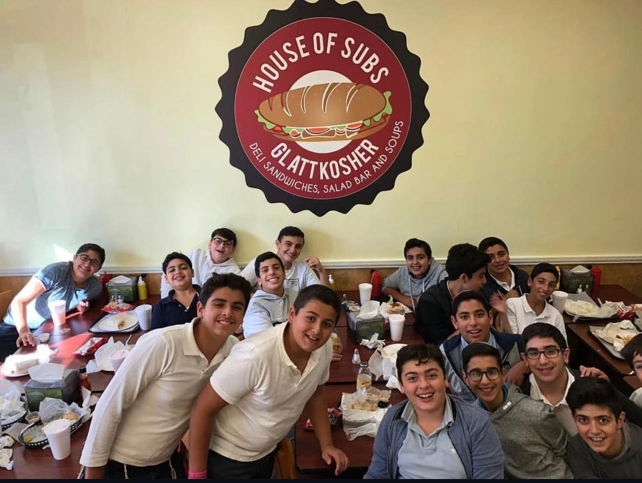 Group of young boys sitting at a long table in a restaurant, smiling, with food and drinks in front of them, in a restaurant called 'House of Subs' which features a large logo of a sandwich on the wall.