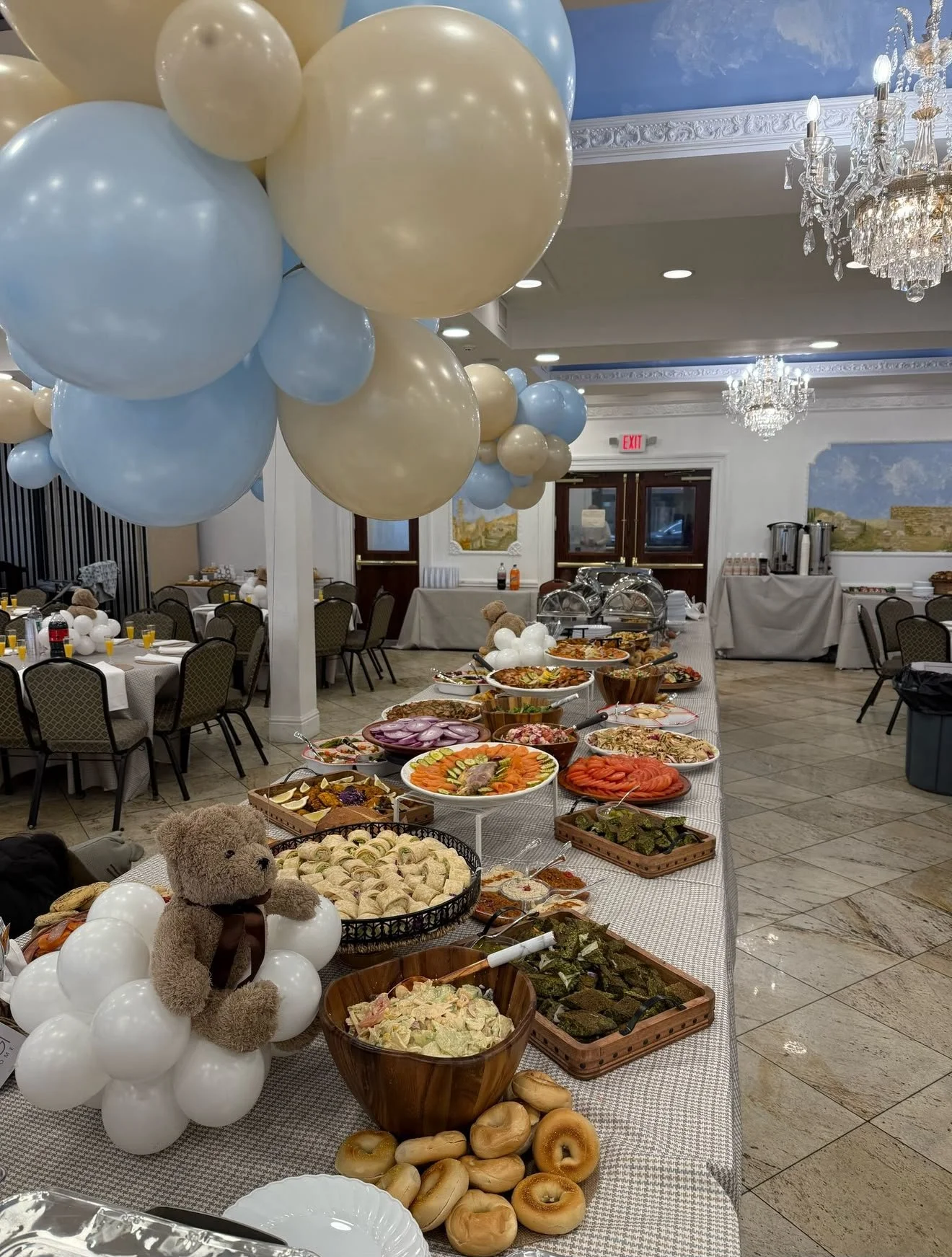 Buffet table with various dishes including salads, vegetables, and finger foods decorated with white and light blue balloons in a decorated dining hall with chandeliers.