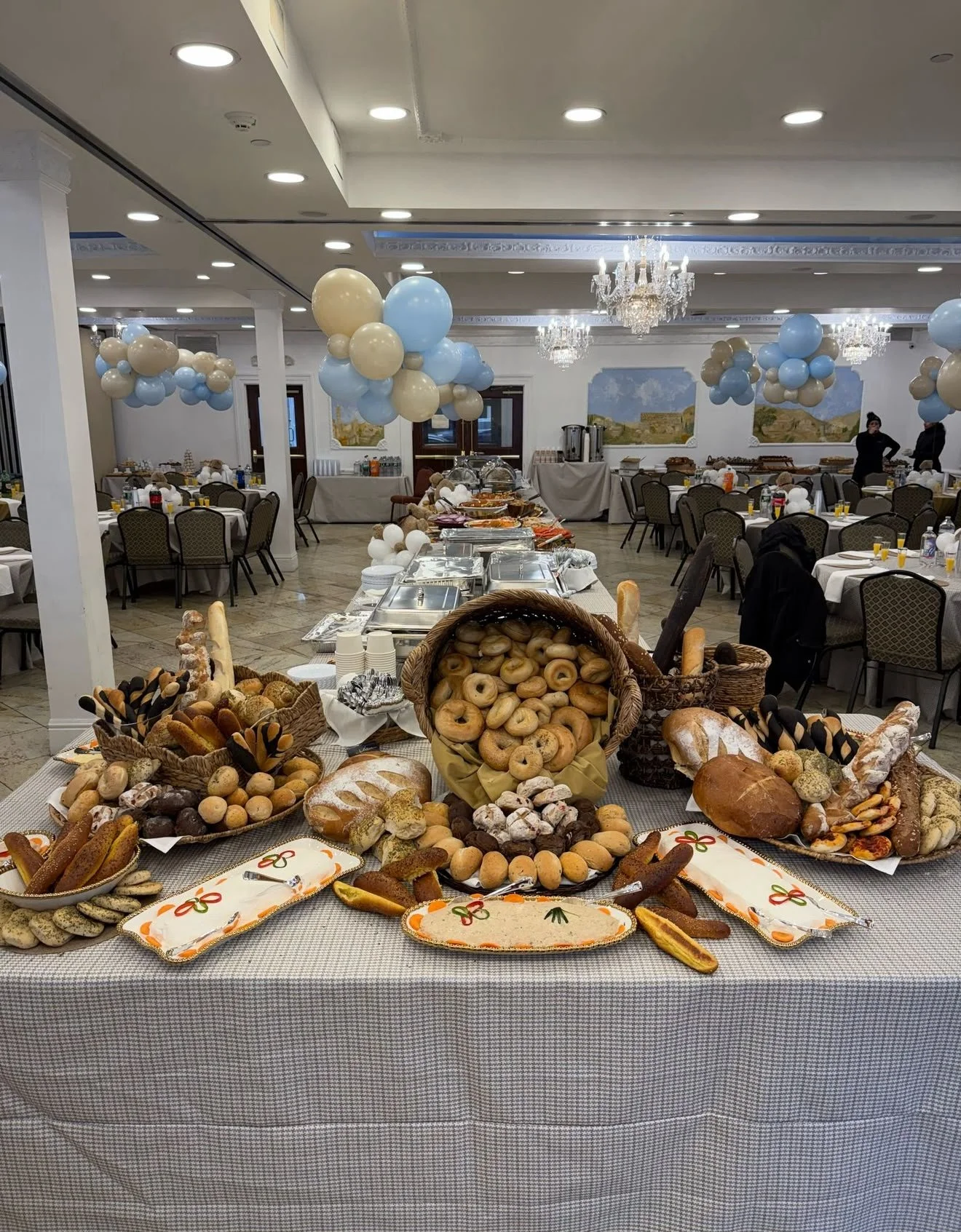 A large buffet table with a variety of breads and baked goods at an event, decorated with balloons in the background and arranged in a banquet hall.