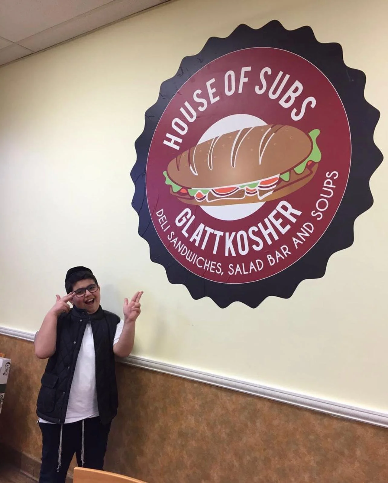 A boy smiling and making a peace sign inside a restaurant with a large circular sign that reads 'House of Subs Glattkosher, Deli Sandwiches, Salad Bar and Soups' on the wall behind him.
