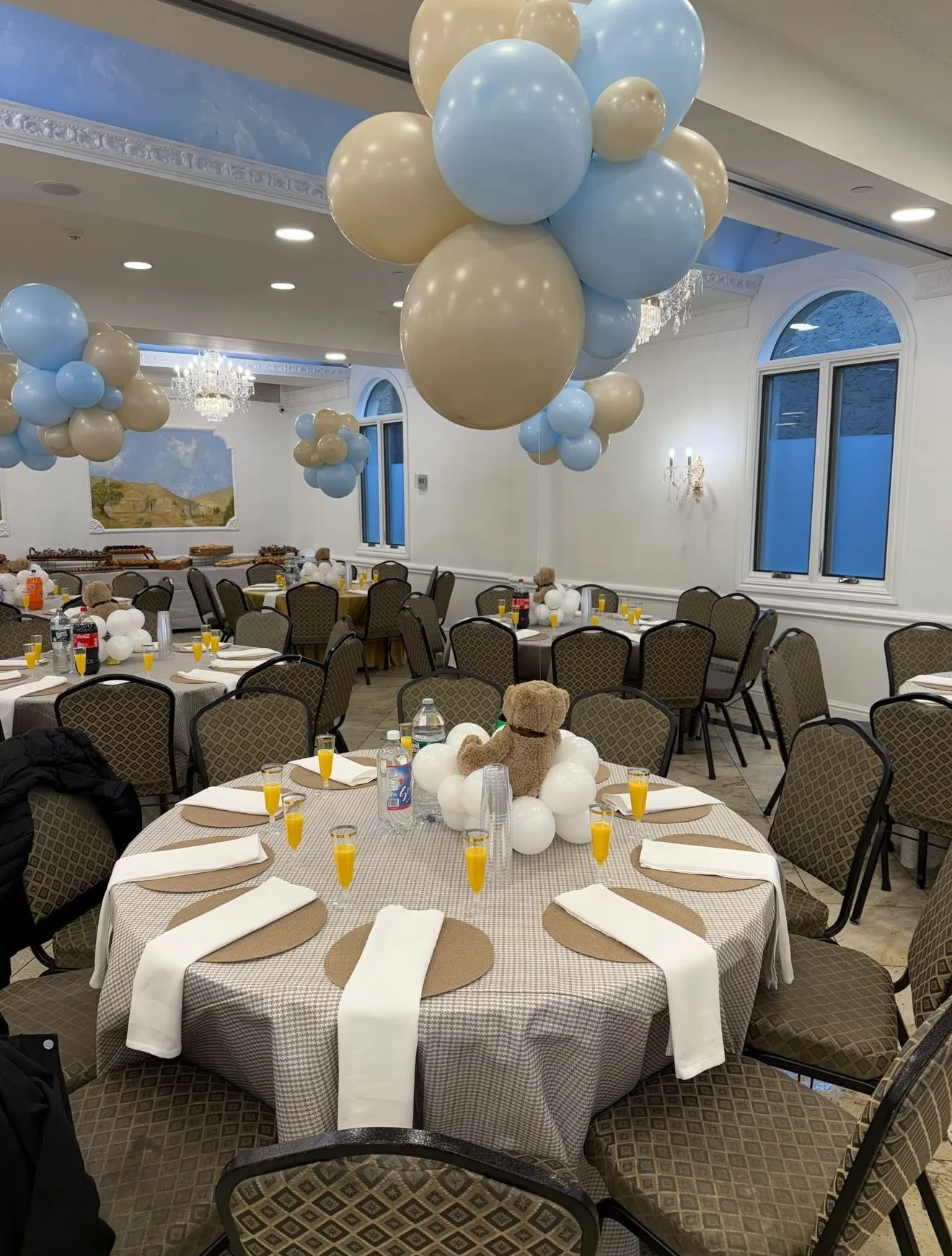 Event hall decorated with blue and beige balloons hanging from the ceiling, round tables with beige and white tablecloths, teddy bears surrounded by white balloons as centerpieces, and chairs set for a celebration.