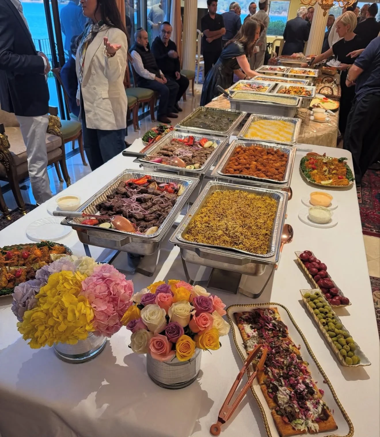 A buffet table with various dishes, including rice, vegetables, meats, and desserts, with guests serving themselves and socializing in a decorated dining area.