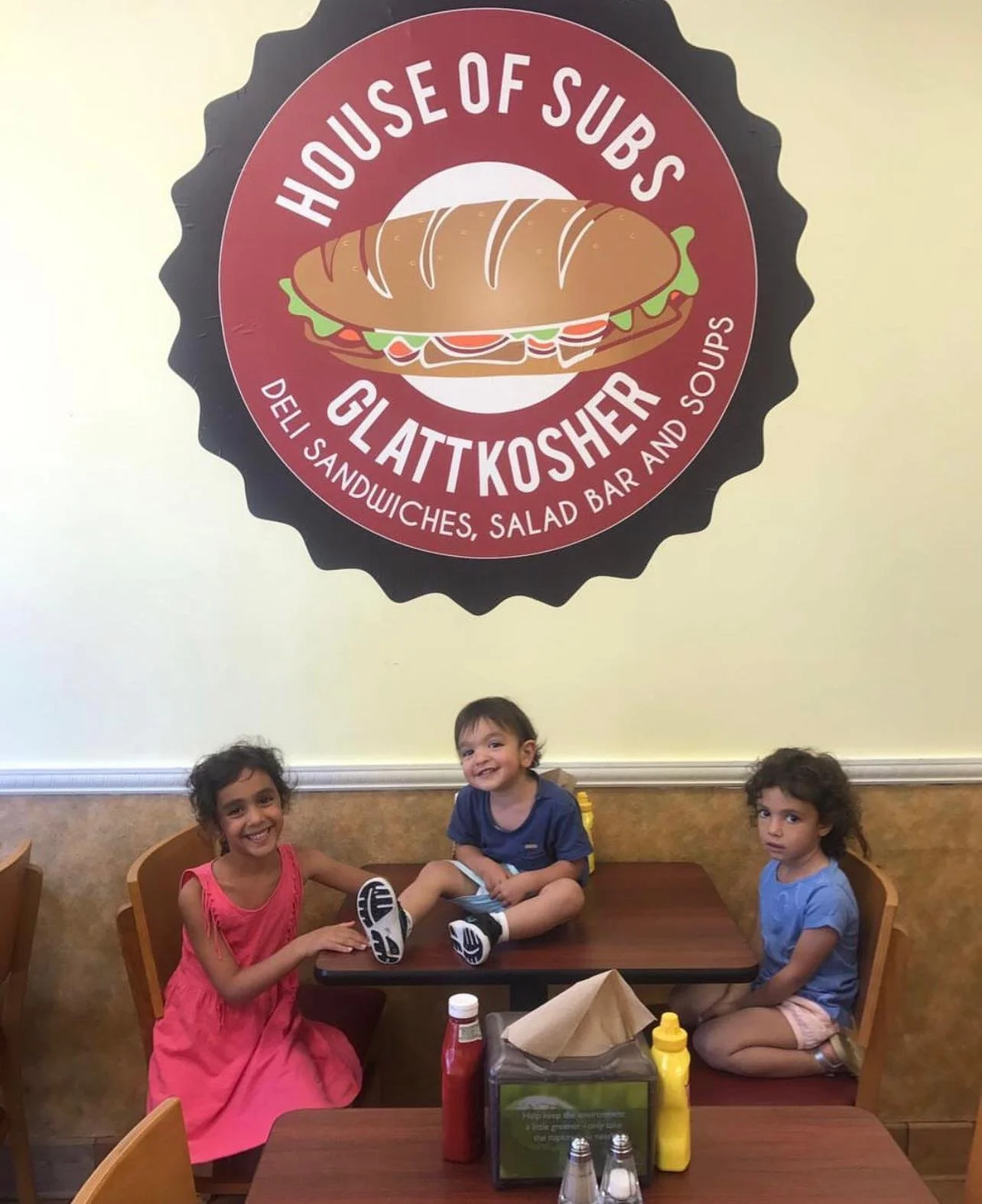 Three children sitting at a table in a restaurant called 'House of Subs Glatt Kosher,' with a sign on the wall featuring a sandwich and the restaurant's name.
