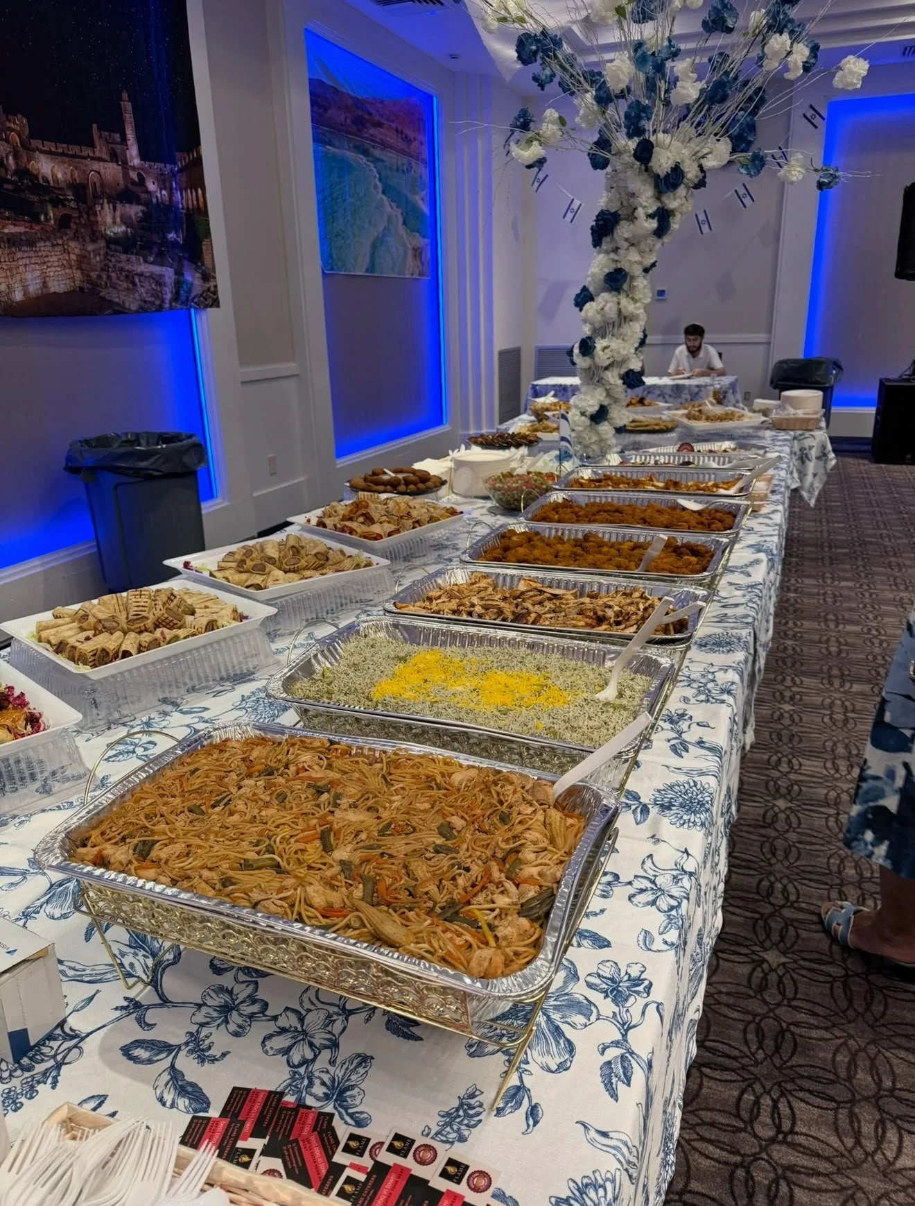 Buffet table with various dishes including pasta, fried chicken, and cookies. A large floral centerpiece stands in the middle of the table. Background features blue lighting, wall art, and a person seated at a table.