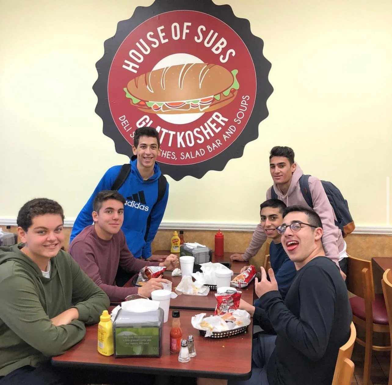 A group of six boys sitting at a table in a restaurant, with two standing behind them, all smiling and posing for a photo. The restaurant wall in the background has a large sign that reads 'House of Subs GLUTT KOSHER' with a sandwich illustration.