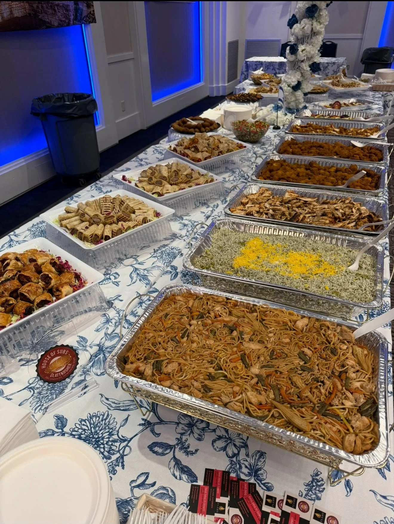 Buffet table with various trays of food including pasta, fried chicken, desserts, and salad at an indoor event with decorative flowers and blue lighting.