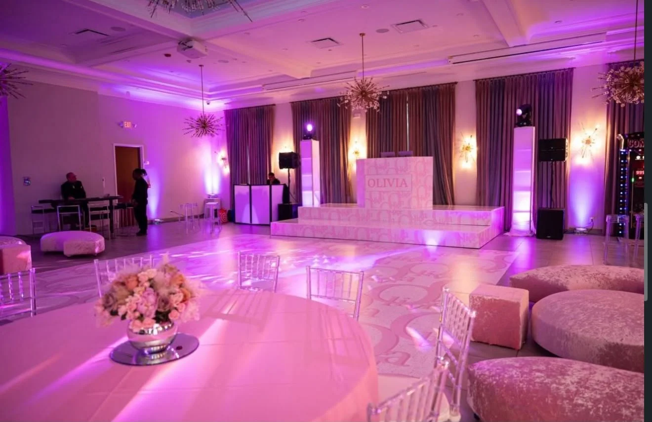 An event hall decorated with pink and purple lighting, featuring a dance floor, a stage with a DJ booth, tables with floral centerpieces, and plush seating.