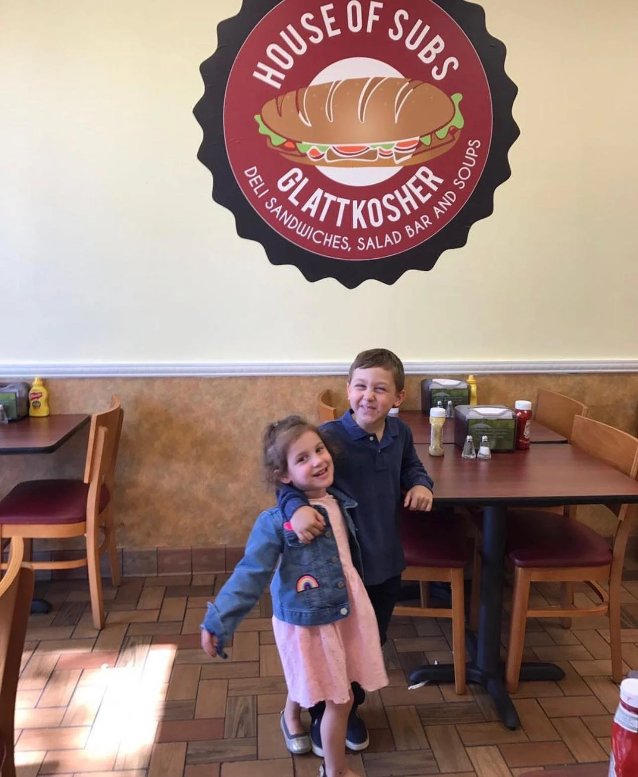 Two children, a girl and a boy, hugging and smiling inside a restaurant with tables, chairs, and condiments on the table. There is a large circular sign on the wall behind them that reads "House of Subs Gluten Kosher" with an image of a sub sandwich.