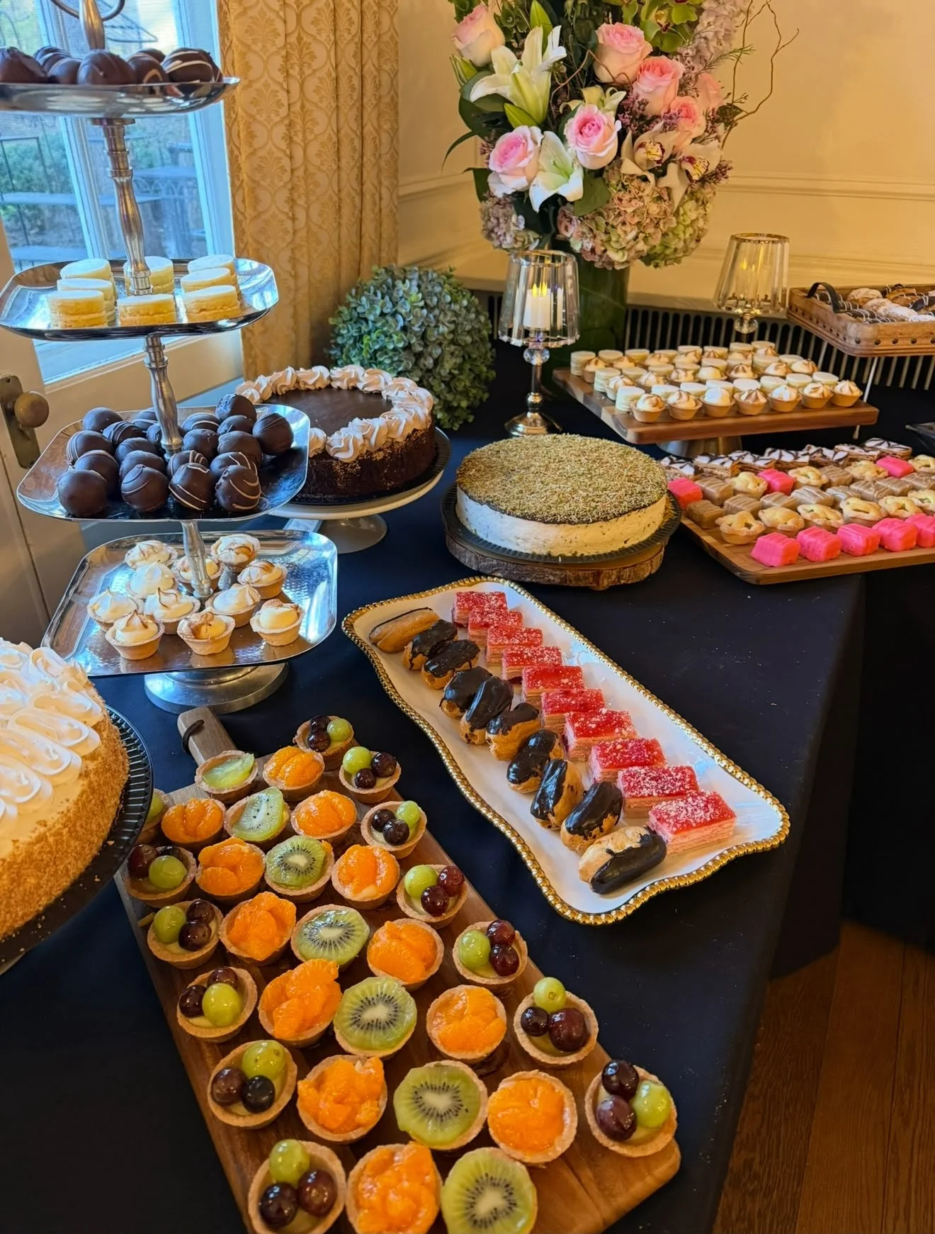 A dessert table featuring an assortment of pastries, cakes, and fruits. There are small fruit tarts with kiwi, grapes, and oranges, a variety of mini eclairs, layered cakes with pink and red frosting, a large round cake with creamy frosting, and an a