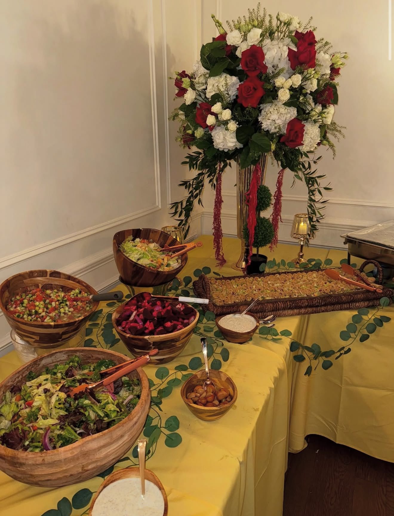 A large floral arrangement with red and white flowers in a gold vase on a decorated table, with bowls of salad, vegetables, and other buffet items around it.