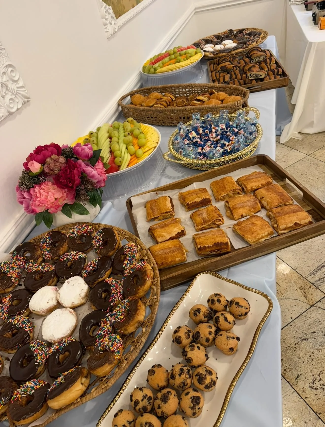 A variety of desserts and snacks arranged on a banquet table, including donuts with colorful sprinkles and icing, chocolate chip cookies, muffins, pastries, fruit platters, and candies, with a floral centerpiece on the side.