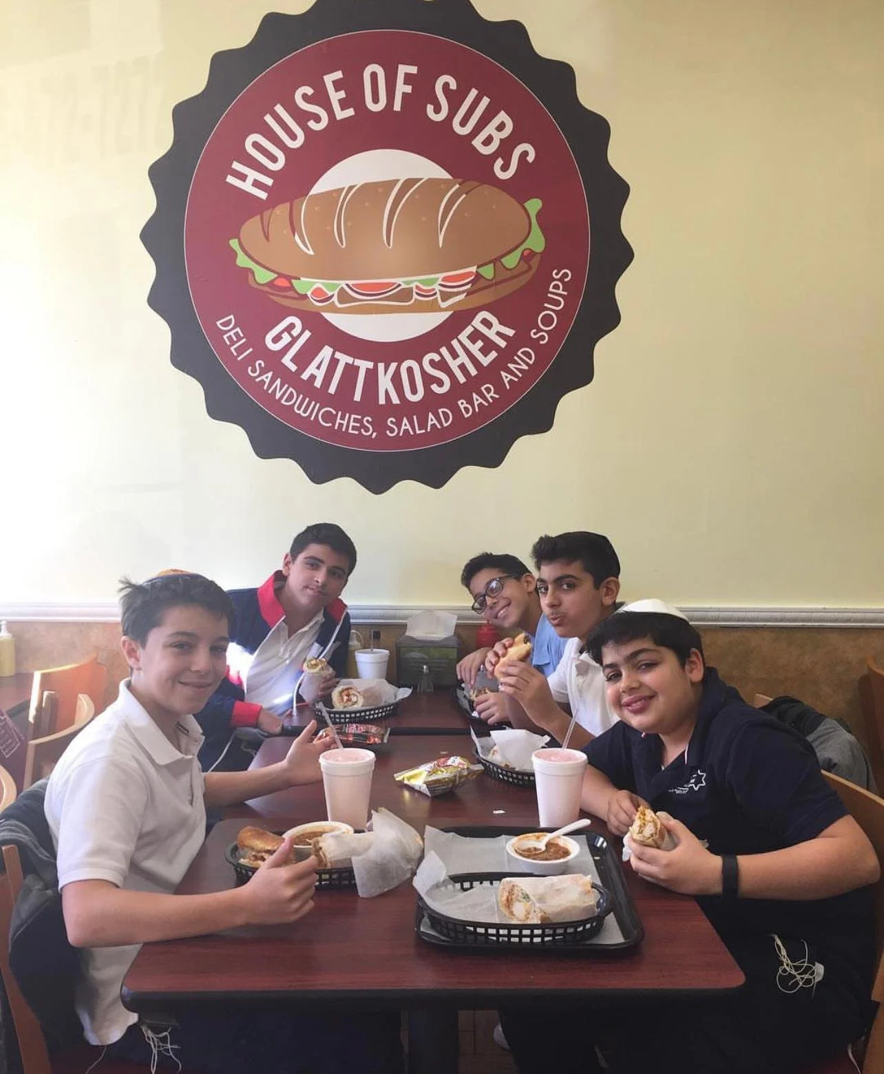 A group of five boys sitting at a restaurant table enjoying sandwiches and drinks, with a large logo for "House of Subs Gluten Kosher" on the wall behind them.