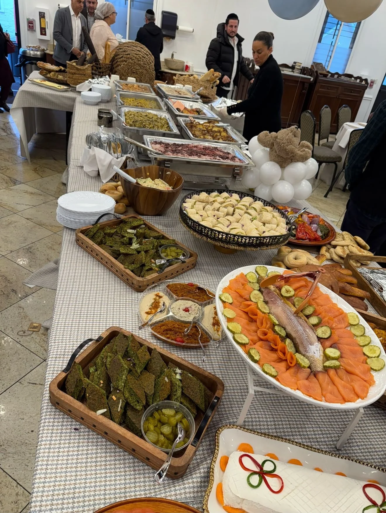 Buffet table with various dishes including smoked fish with cucumber slices, salads, and baked foods, with people serving themselves at a gathering
