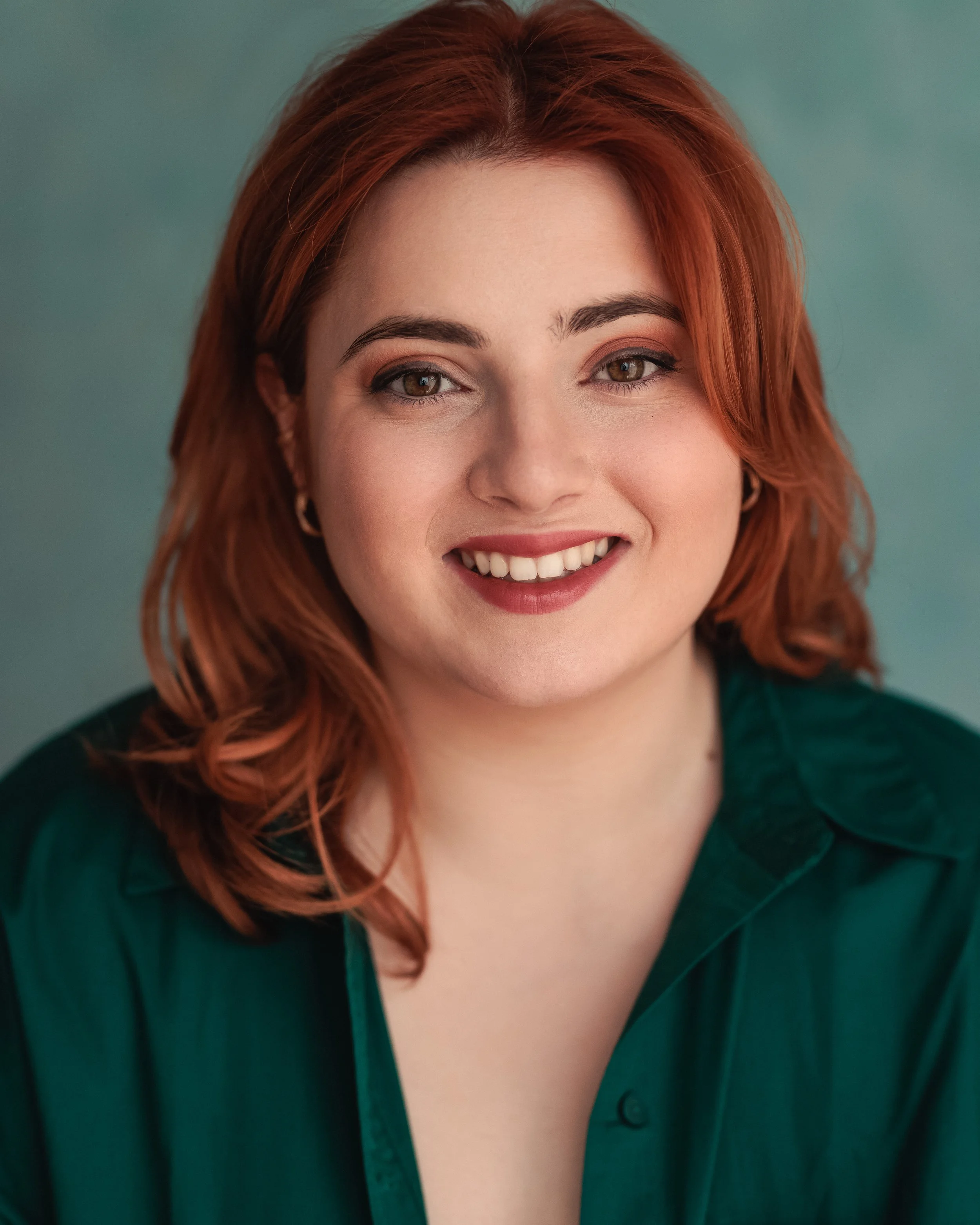 Close-up of a smiling woman with red hair, wearing a dark green top, against a teal background.