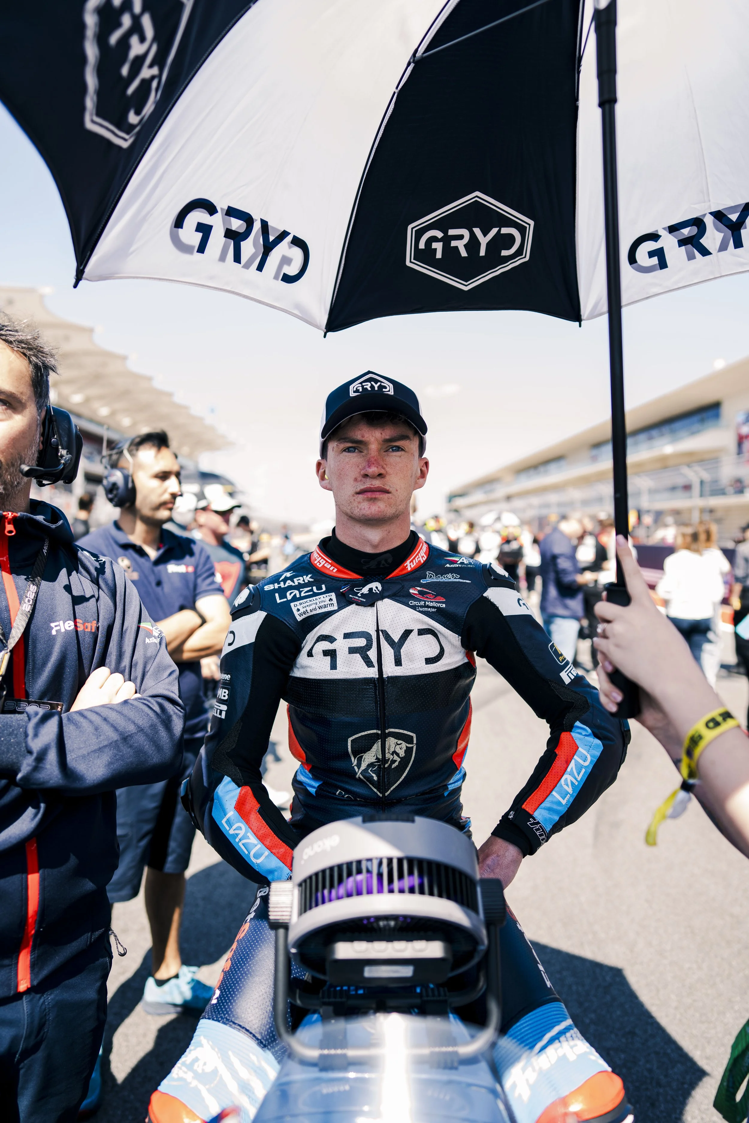 A professional motorcycle racer in racing gear standing at the starting line of a race track, with team members and officials around, holding umbrellas to shield from the sun.
