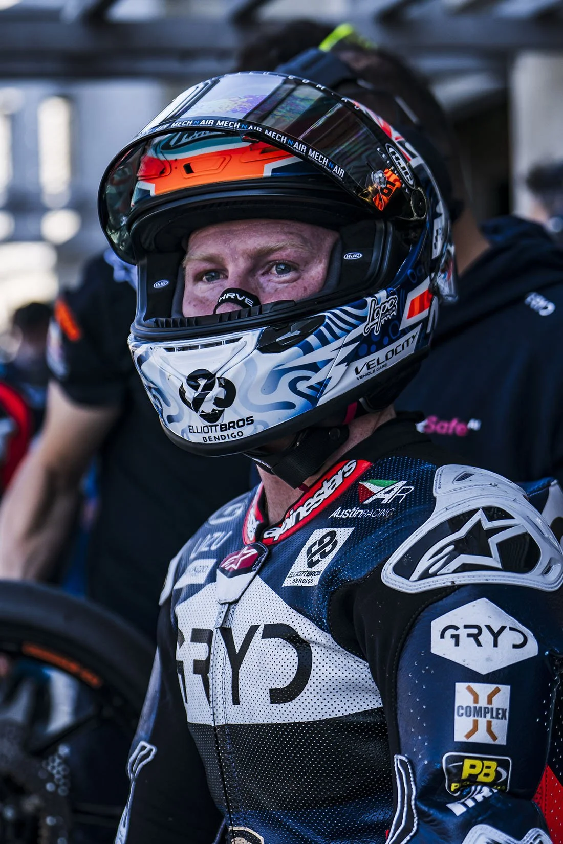 A race car driver wearing a full racing suit and a helmet, with various sponsor logos, looking into the camera.