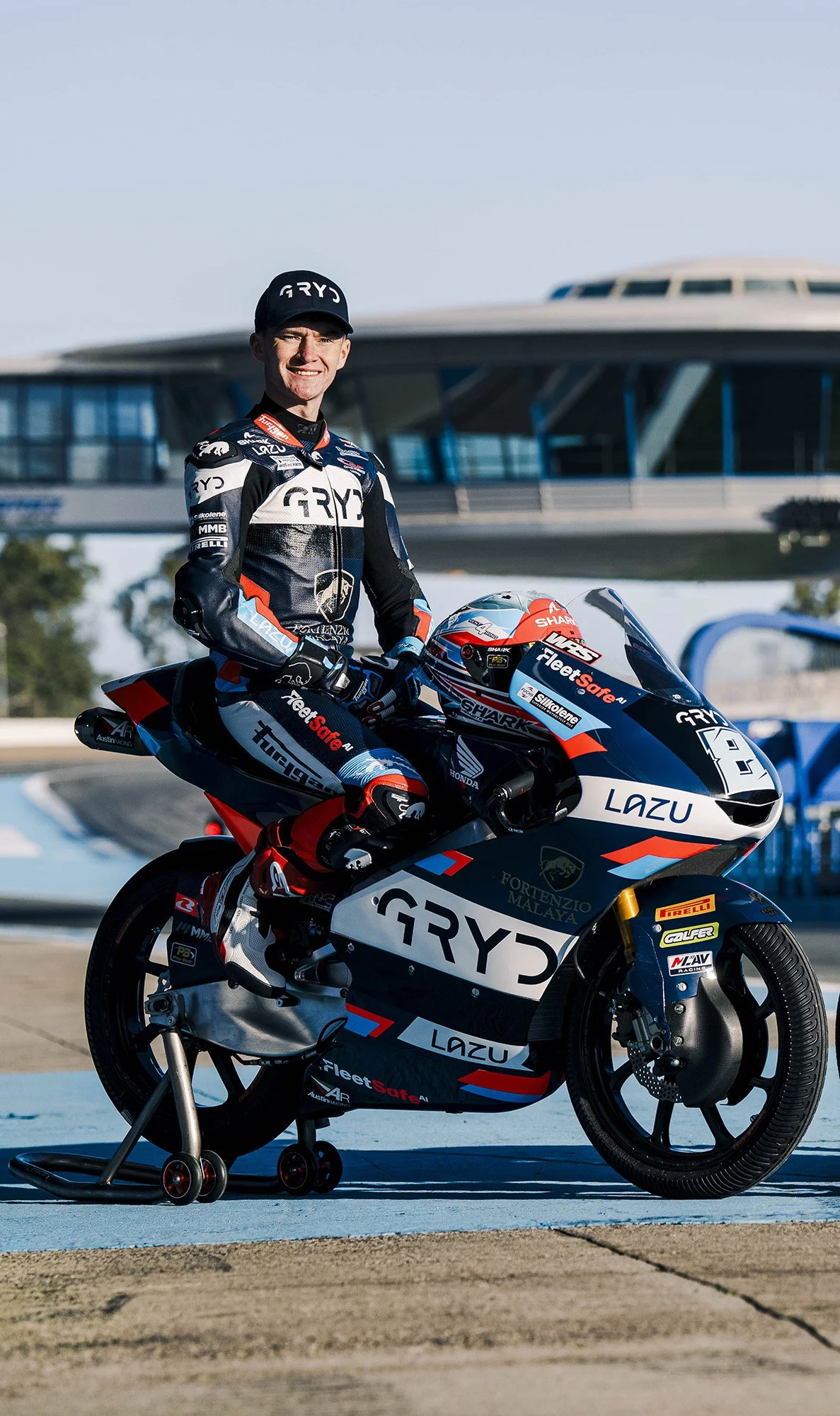 A professional motorcycle racer in racing gear sitting on a race bike at a racetrack, with a large modern building in the background.