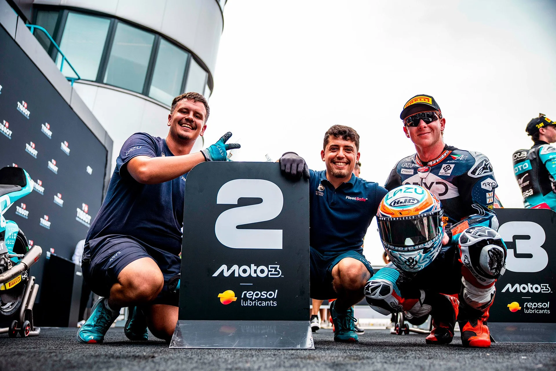 Three men at a motorcycle racing event, kneeling with a podium and racing gear, smiling and posing, with a motorcycle and event branding visible.
