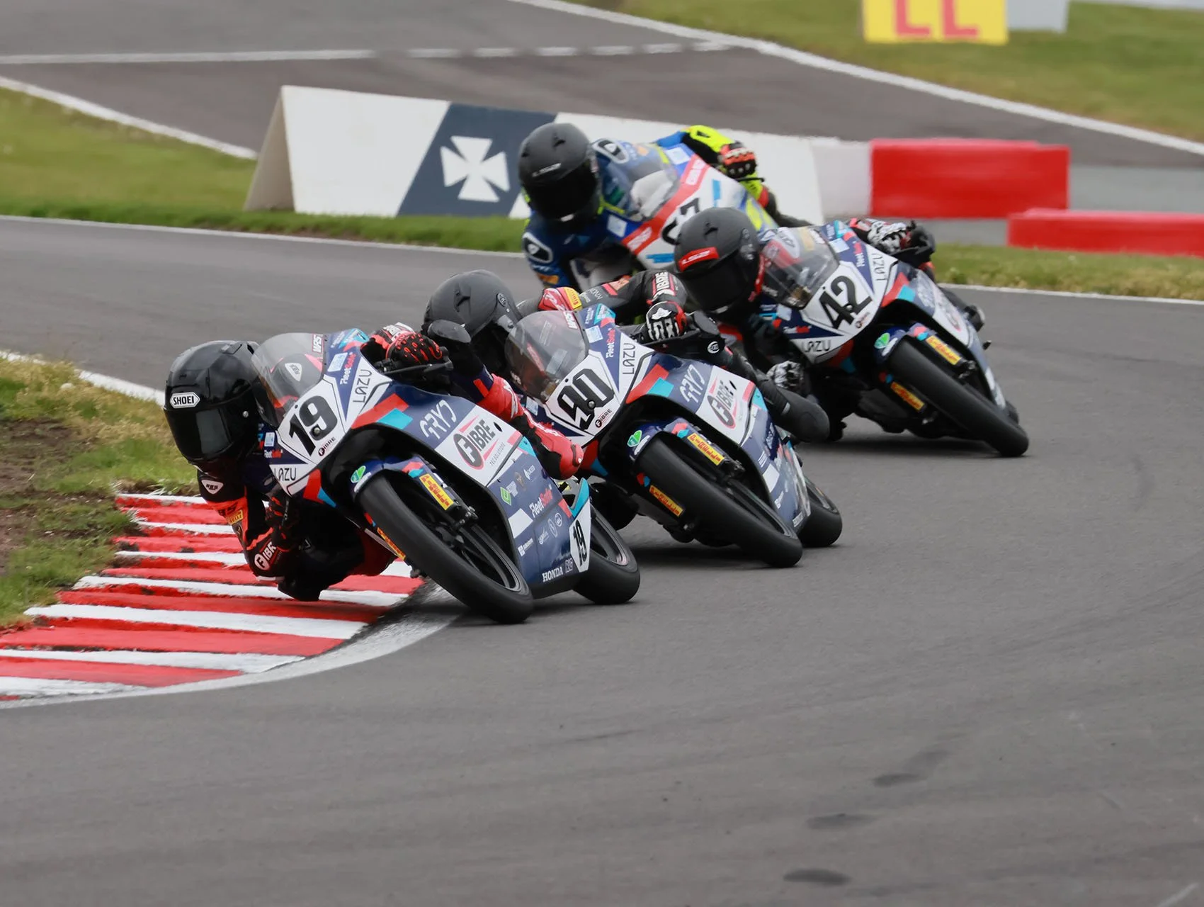 Three motorcycle racers leaning into a curve on a racetrack, with tires close to the red and white striped curb, surrounded by grass and safety barriers.
