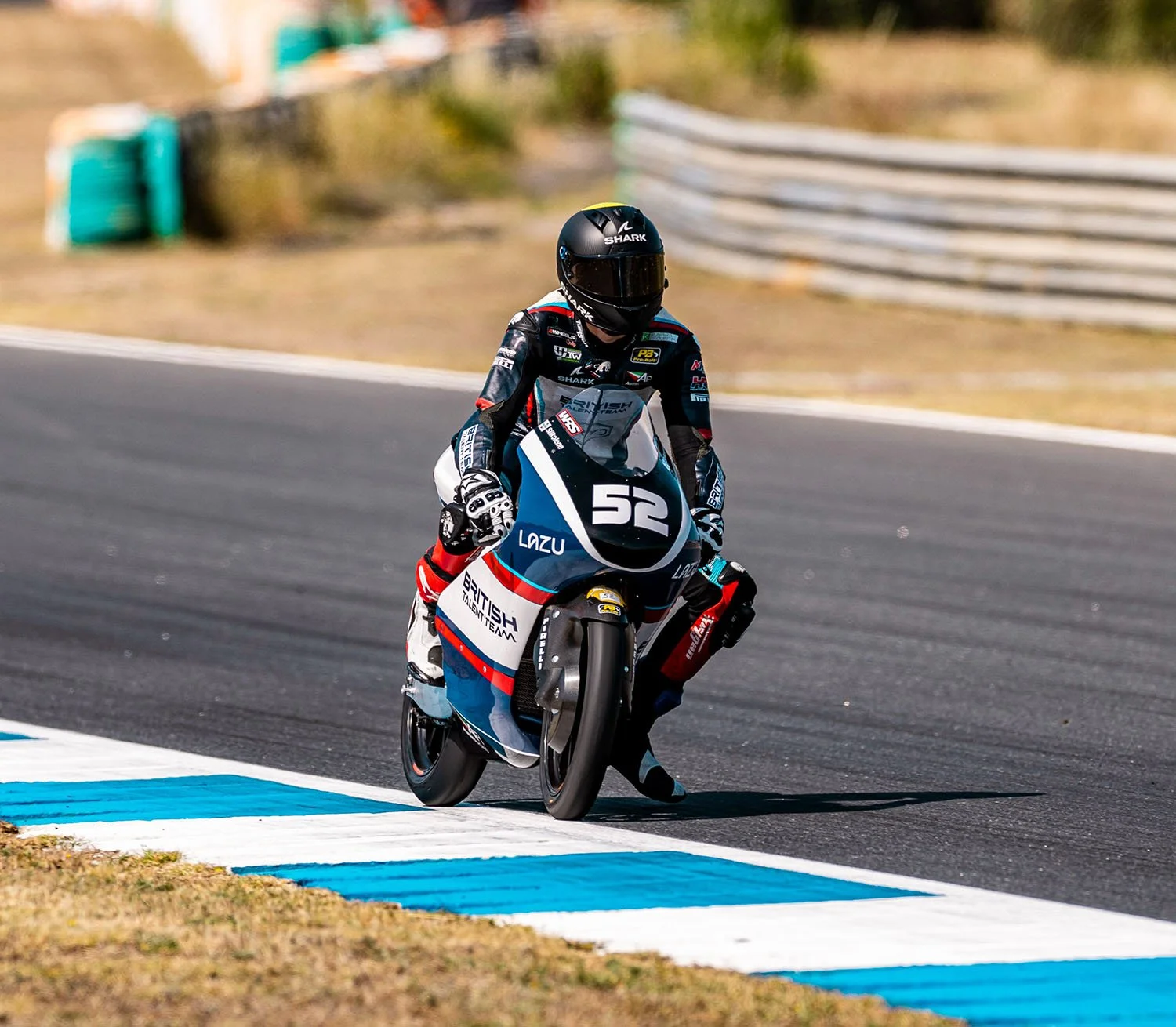Motorcycle race car driver wearing a black helmet, black and blue racing suit, riding a racing bike numbered 52 on a racetrack, with blurred background.