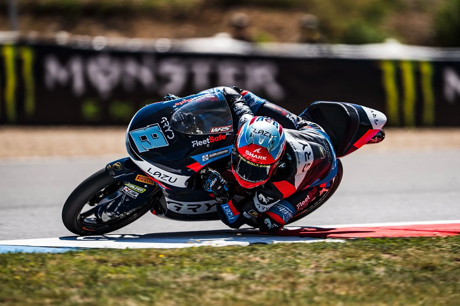 Motorcycle racer leaning into a turn on a race track, dressed in racing suit and helmet, with sponsor logos visible on the bike and gear.