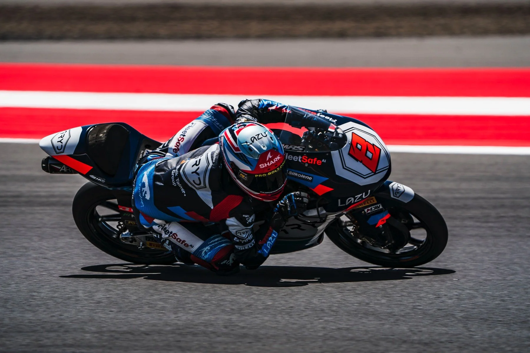 Motorcycle racer leaning into a turn on a race track, wearing a black, blue, and white racing suit and helmet with number 86 on the bike, with red and white lines on the track surface.