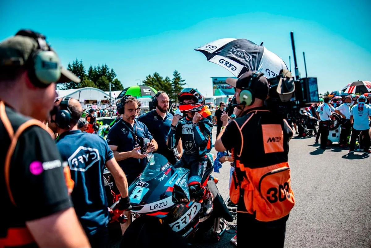 Motorcycle racer in racing suit adjusting helmet while surrounded by team members and camera crew at a racetrack during daytime.
