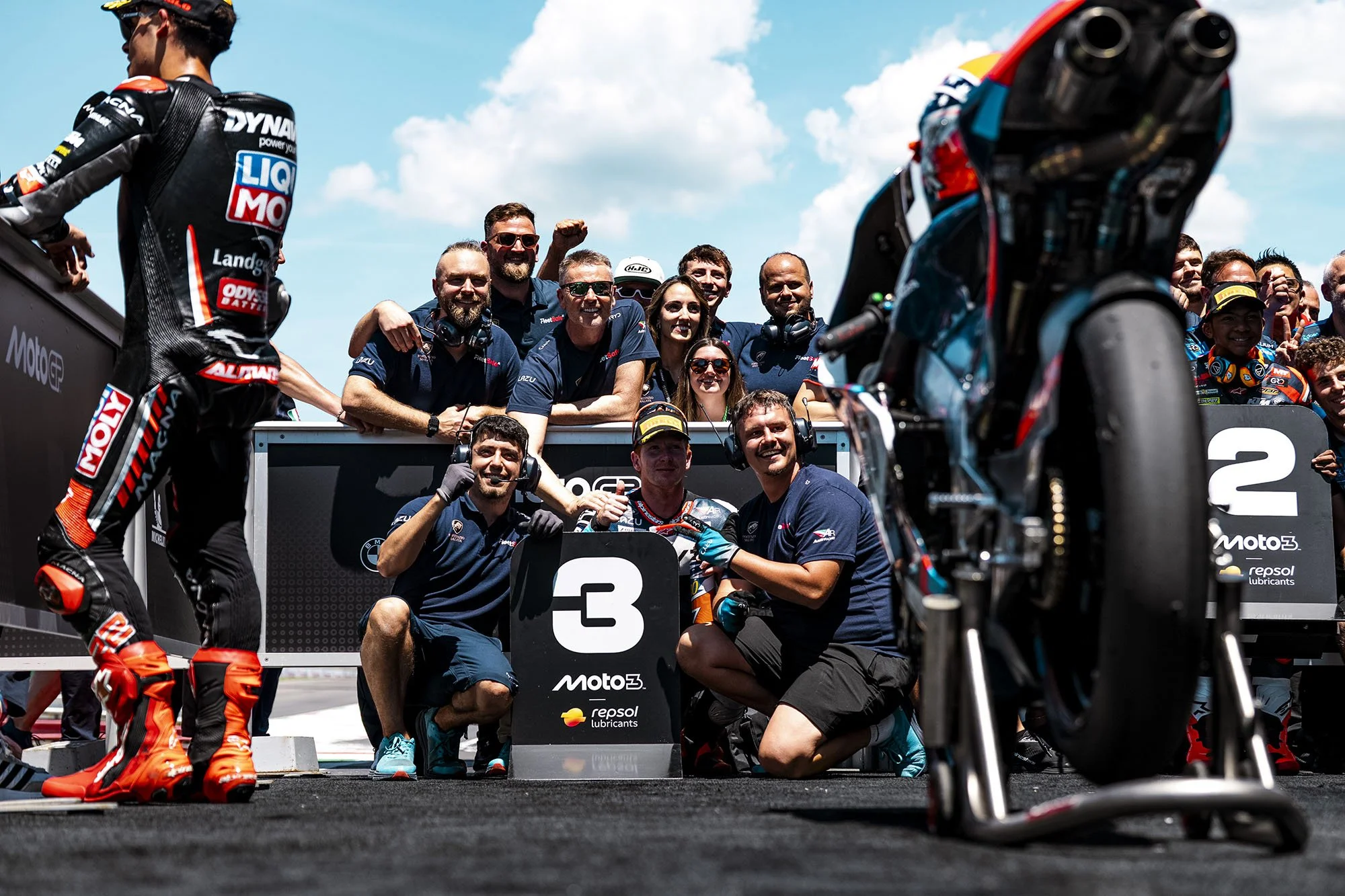 Motorcycle racing team celebrating on the podium with race winners, teams, and mechanics, with a Yamaha motorcycle in the foreground during a motorsport event.
