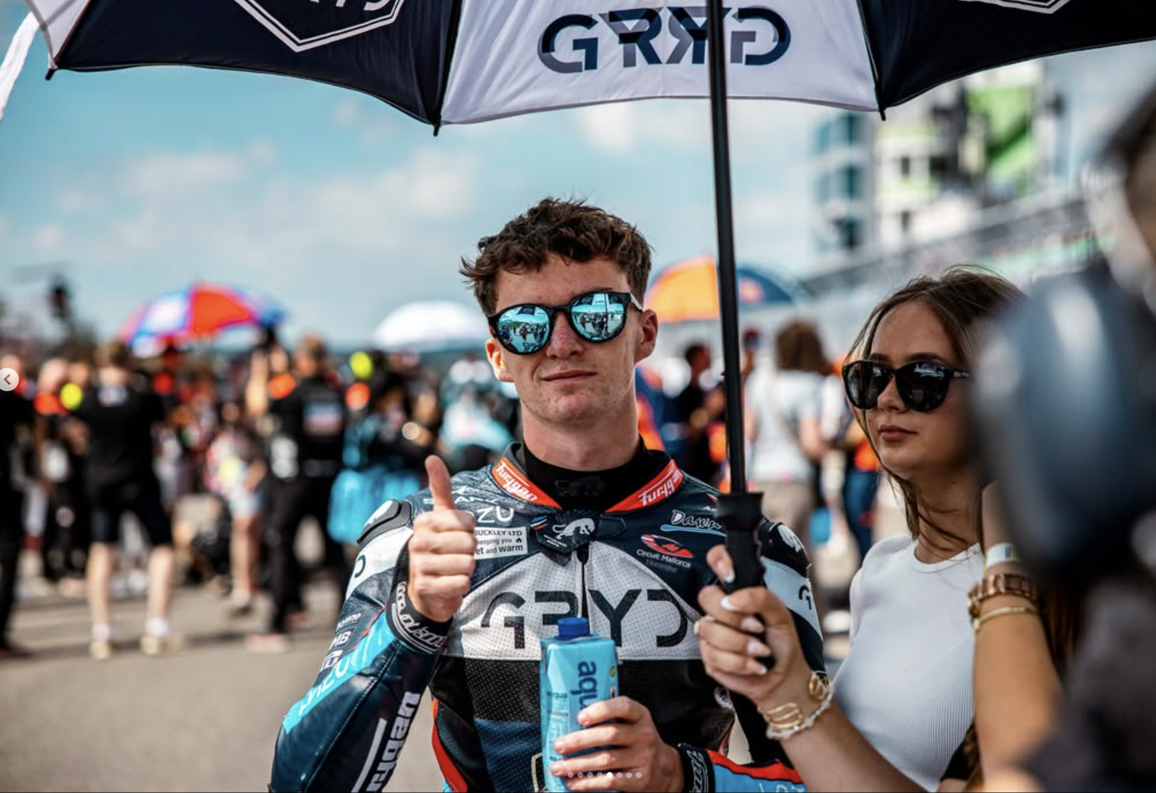 A man in racing gear holding a water bottle, giving a thumbs-up, under an umbrella with the word 'GRYD' on it, surrounded by a crowd at a racing event.
