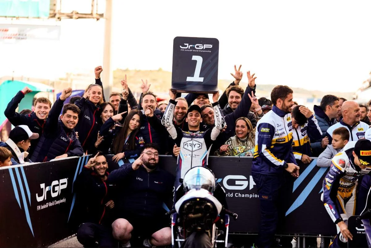 Motorsport team celebrating victory at the FIM JuniorGP World Championship, with one member holding a trophy with a large number 1, surrounded by smiling team members and officials.