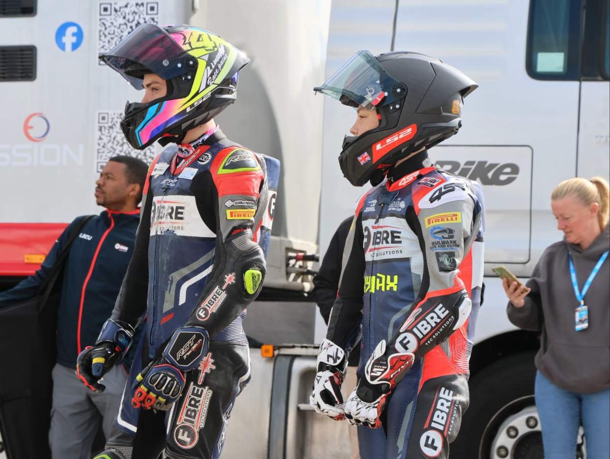 Two motorcycle racers in full gear wearing helmets and racing suits standing next to each other while two women are nearby, one looking at a phone and another standing in the background.