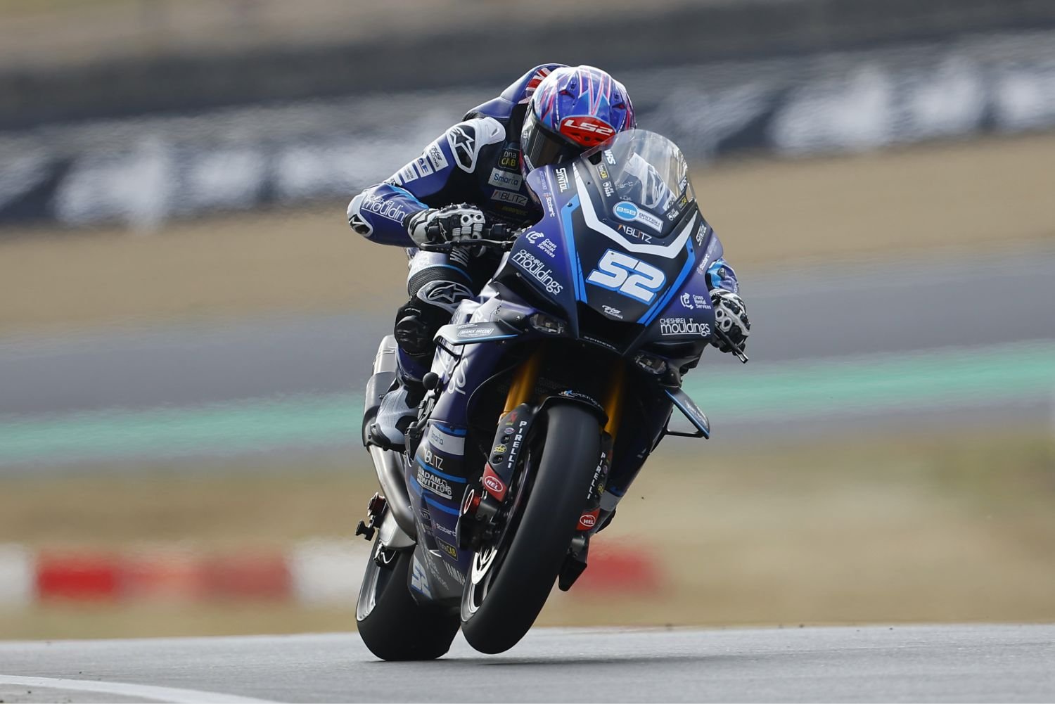 A motorcycle racer wearing a blue and black racing suit and helmet is leaning into a turn on a race track, riding a blue racing motorcycle with the number 52 on it.