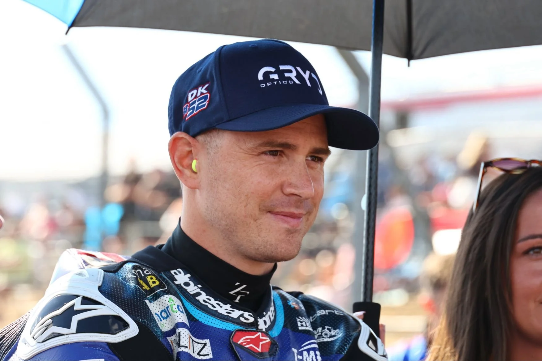 Close-up of professional motorcycle racer wearing racing suit and cap, standing under umbrella, with blurred background of cars and spectators at a racetrack.