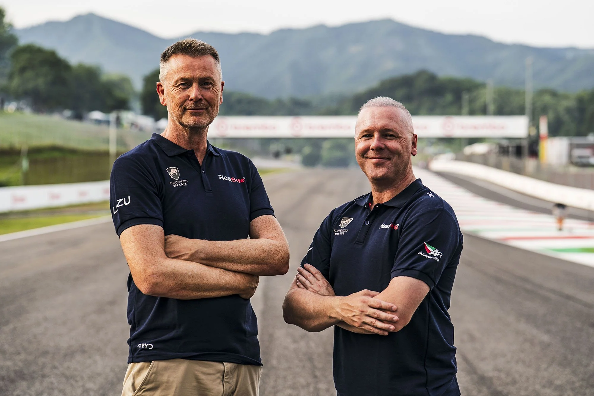 Two men in navy blue shirts with sponsor logos standing on a racing track with mountains in the background. They have crossed arms and are smiling.