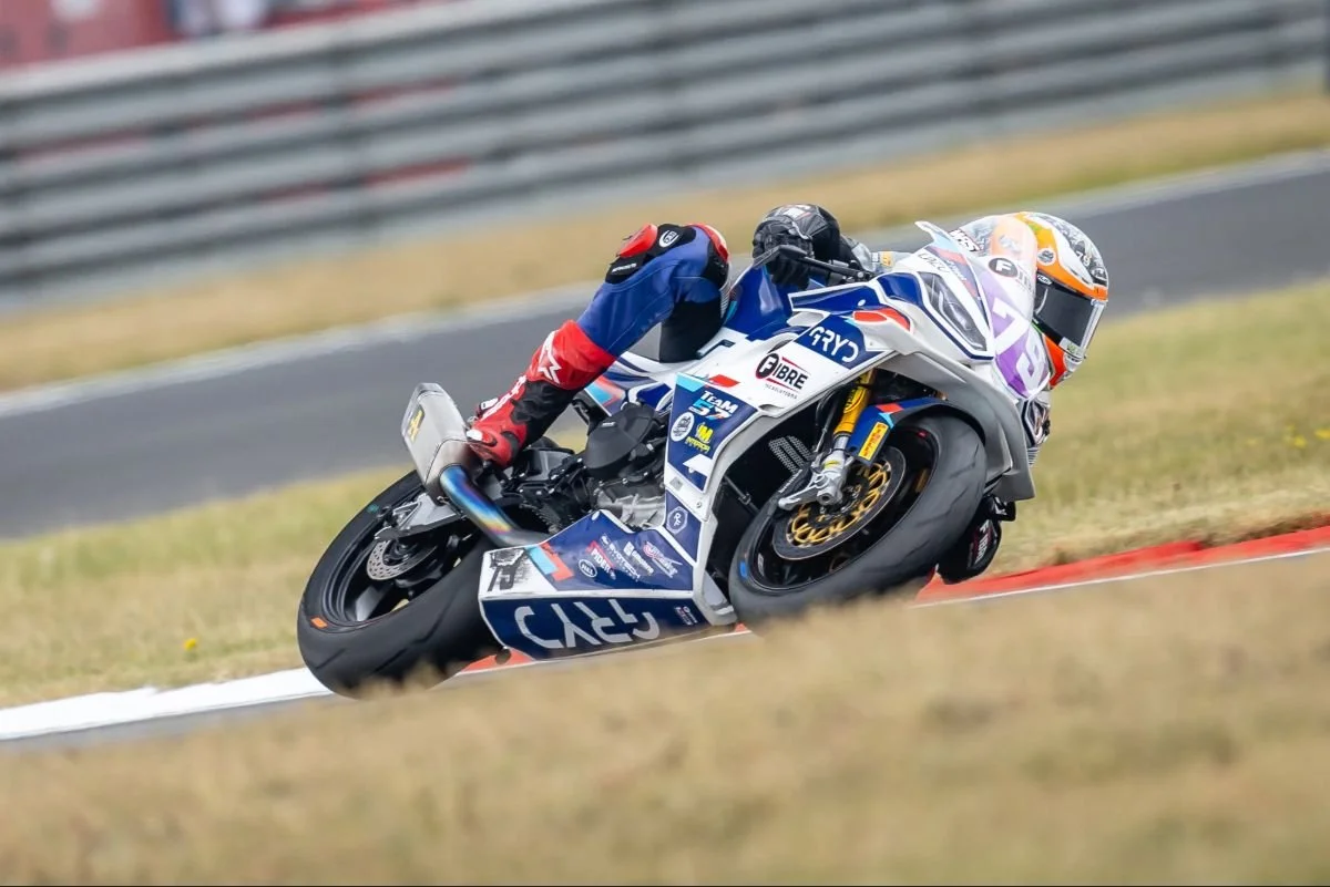 Motorcycle racer leaning into a turn on a race track, wearing a racing suit and helmet, with sponsor logos on the bike and suit.