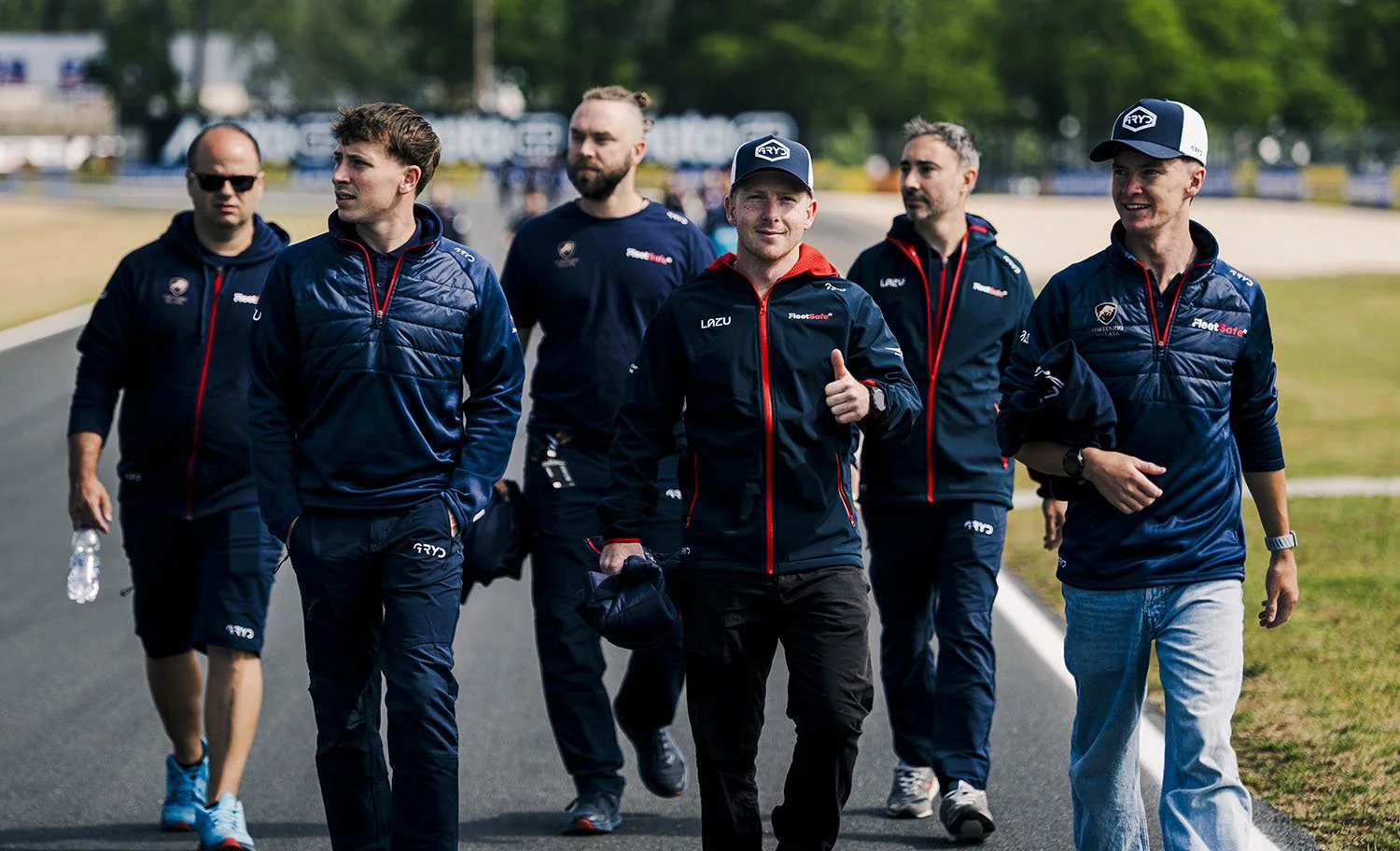 Group of race car drivers and team members walking on a track, wearing team uniforms with logos and some wearing caps, with a grassy area and trees in the background.