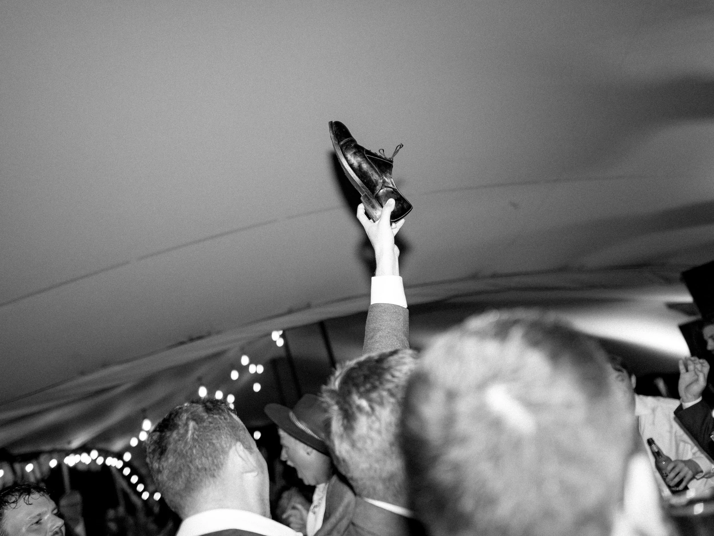 A person raises a worn leather shoe into the air at a crowded event, with a ceiling featuring string lights in the background.