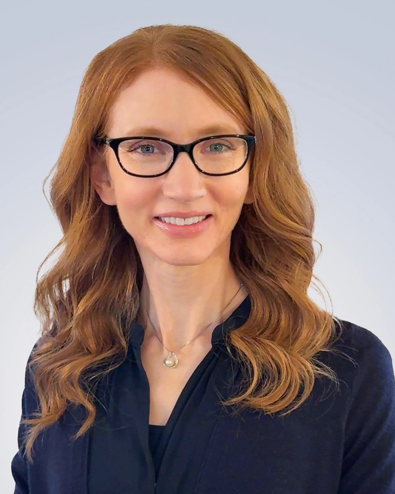Portrait of Christine Dotzenrod Childress, MS, CCC-SLP with long red hair, glasses, and a necklace, wearing a dark top, smiling in front of a light background.